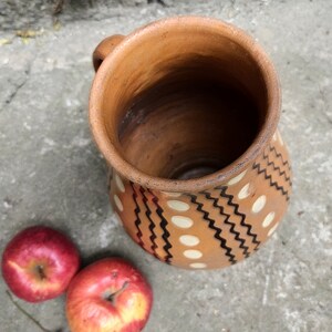 May include: A handmade, terracotta-colored ceramic pitcher with a handle, decorated with white dots and black zigzag patterns. Two red apples are next to the pitcher, offering a rustic, natural aesthetic.