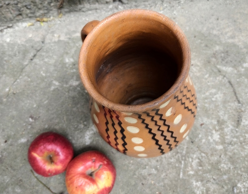 May include: A handmade, terracotta-colored ceramic pitcher with a handle, decorated with white dots and black zigzag patterns. Two red apples are next to the pitcher, offering a rustic, natural aesthetic.