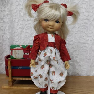 Peut inclure: Une poupée aux cheveux blonds en couettes portant une veste rouge et une robe blanche à motif brun et blanc. La poupée est debout sur un support blanc.
