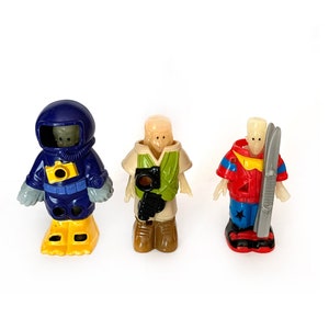 May include: Three plastic figurines, each with a different theme. The first is a blue astronaut with a yellow camera, the second is a green explorer with a brown camera, and the third is a red skier with a white ski.