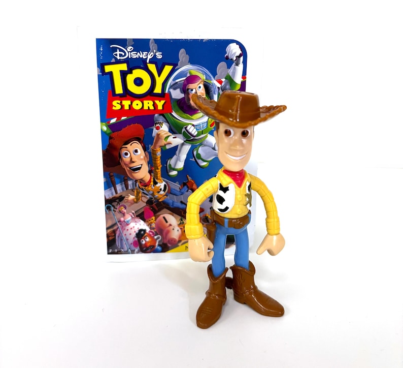 May include: A plastic toy figure of Woody from the Disney Pixar Toy Story franchise. Woody is wearing his signature brown cowboy hat, a yellow vest with a red bandana, and blue jeans. He is holding a sheriff's badge and has a friendly smile. The figure is standing in front of a Toy Story movie packaging.