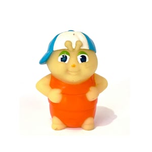 Puede incluir: Una pequeña figura de goma de color claro que representa un personaje de dibujos animados con grandes ojos verdes y dos pequeñas antenas en la cabeza. El personaje lleva una gorra de béisbol azul y blanca y un traje naranja segmentado.