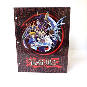Puede incluir: Un archivador Yu-Gi-Oh! negro y rojo con personajes de la serie de anime. El archivador tiene una imagen central de varios personajes y monstruos, con el logotipo de Yu-Gi-Oh! en la parte inferior. La portada está adornada con patrones de estilo jeroglífico.