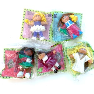 Puede incluir: Cinco muñecas Cabbage Patch Kids en su embalaje original. Cada muñeca está en una bolsa de plástico transparente con un respaldo estampado en verde y blanco. Las muñecas tienen varios atuendos y peinados, incluyendo cabello rubio, castaño y negro.