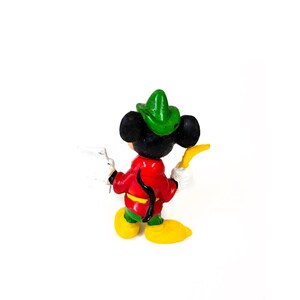 May include: A small figurine of a black mouse character wearing a green hat, a red jacket with a yellow belt, green pants, and yellow shoes. The character holds a white object in one hand and a curved yellow object in the other hand.