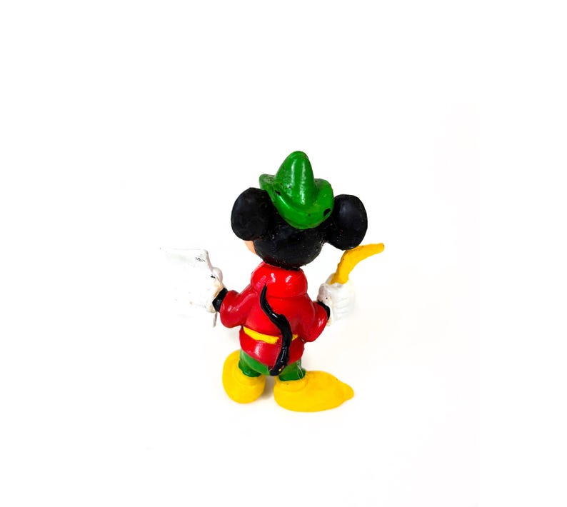 May include: A small figurine of a black mouse character wearing a green hat, a red jacket with a yellow belt, green pants, and yellow shoes. The character holds a white object in one hand and a curved yellow object in the other hand.