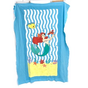 May include: A light blue beach towel featuring a cartoon mermaid with red hair and a green tail. The mermaid is surrounded by sea creatures, including a yellow fish and a red crab, against a backdrop of blue and white stripes.