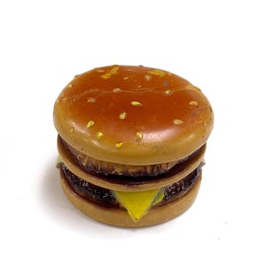 May include: A small, round, burger-shaped object with a brown bun top featuring light-colored sesame seed details. The middle section shows a dark brown patty layer, a yellow triangular cheese slice, and a bottom bun, all with a glossy finish.