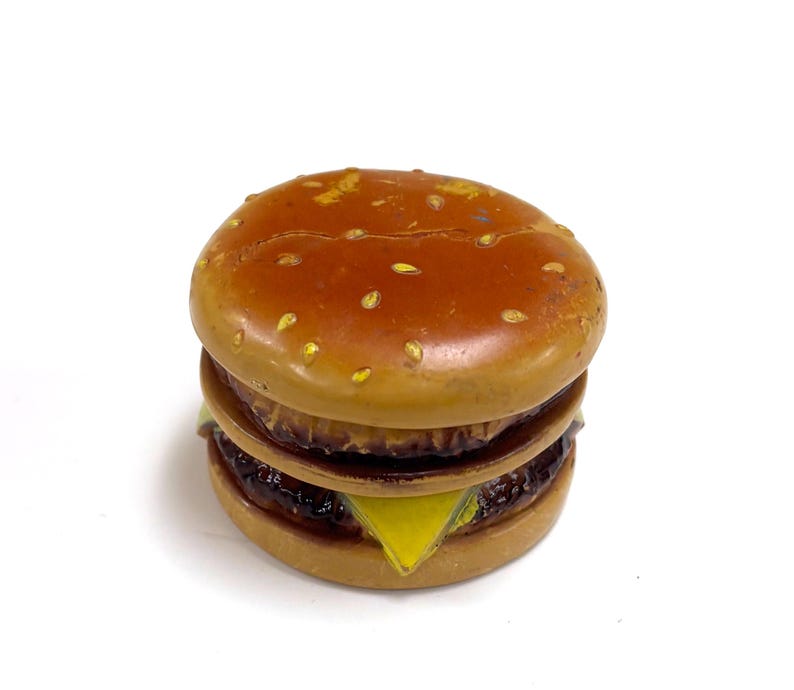 May include: A small, round, burger-shaped object with a brown bun top featuring light-coloured sesame seed details. The middle section shows a dark brown patty layer, a yellow triangular cheese slice, and a bottom bun, all with a glossy finish.