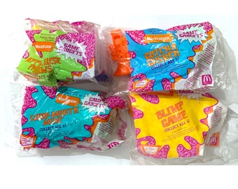 May include: Four Nickelodeon-themed Happy Meal toys from McDonald's. The toys are packaged in clear plastic bags with colourful graphics. The toys include a Loud Mouth Mike, a Gotcha Gasket, a Blimp Game, and a toy gun.