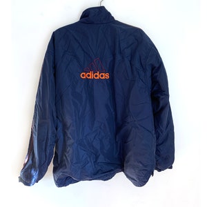 K&ouml;nnte beinhalten: Eine marineblaue Windbreaker-Jacke mit einem roten und orangen Adidas-Logo auf der R&uuml;ckseite.