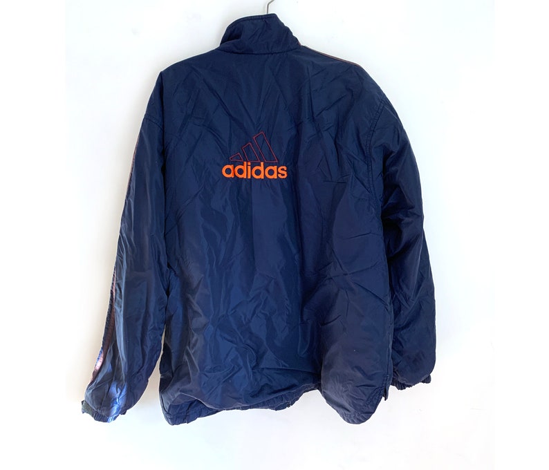 K&ouml;nnte beinhalten: Eine marineblaue Windbreaker-Jacke mit einem roten und orangen Adidas-Logo auf der R&uuml;ckseite.