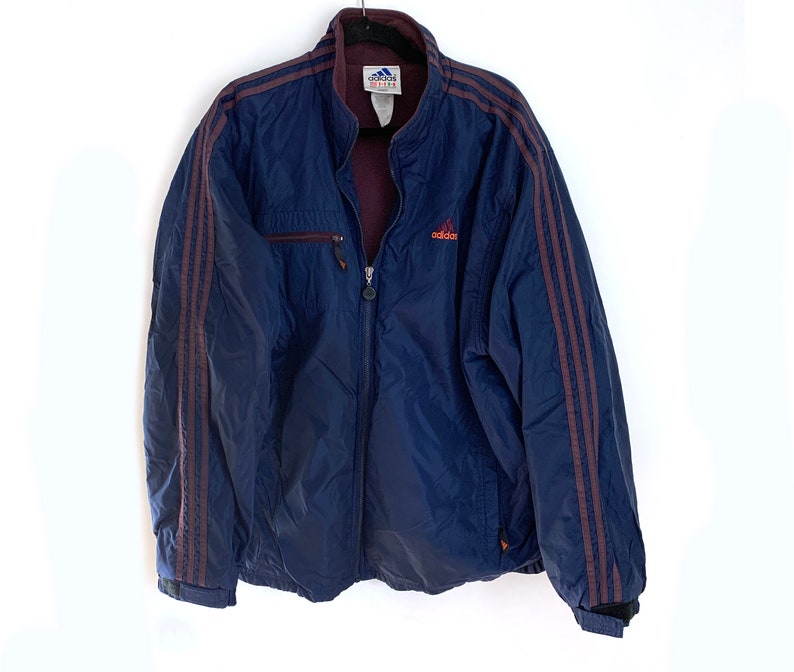 K&ouml;nnte beinhalten: Eine dunkelblaue Windbreaker-Jacke mit maroonfarbenen Streifen an den &Auml;rmeln und einem kleinen orangen "adidas"-Logo auf der Brust.