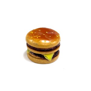 May include: A miniature toy hamburger featuring a glossy brown bun with yellow sesame seed details, a dark brown patty, and a slice of bright yellow cheese visible between the layers. The object has a smooth, rounded, and stacked construction.