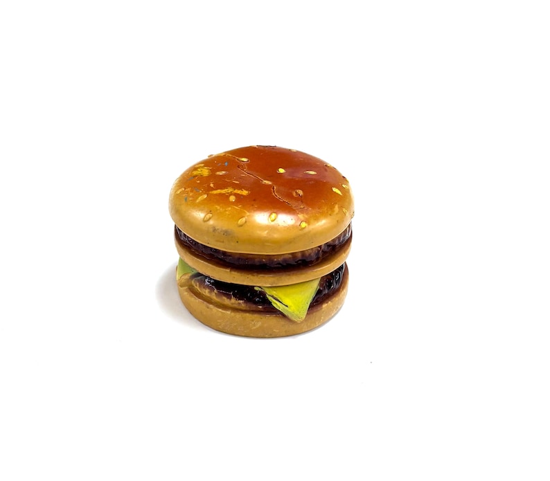 May include: A miniature toy hamburger featuring a glossy brown bun with yellow sesame seed details, a dark brown patty, and a slice of bright yellow cheese visible between the layers. The object has a smooth, rounded, and stacked construction.