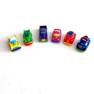 Puede incluir: Una colección de seis coches de juguete de colores. Los coches están hechos de plástico y tienen diferentes diseños. Los coches están todos en fila sobre una superficie blanca.