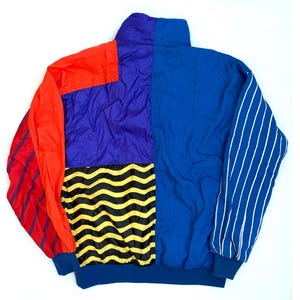 K&ouml;nnte beinhalten: Eine Color-Block-Windjacke mit hohem Kragen und langen &Auml;rmeln. Das Design umfasst einfarbige Paneele in Rot, Lila und Blau, ein schwarz-gelbes Wellenmuster am unteren R&uuml;cken sowie wei&szlig;e vertikale Nadelstreifen auf einem der &Auml;rmel.