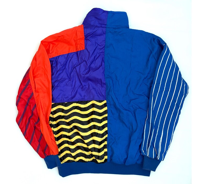 K&ouml;nnte beinhalten: Eine Color-Block-Windjacke mit hohem Kragen und langen &Auml;rmeln. Das Design umfasst einfarbige Paneele in Rot, Lila und Blau, ein schwarz-gelbes Wellenmuster am unteren R&uuml;cken sowie wei&szlig;e vertikale Nadelstreifen auf einem der &Auml;rmel.
