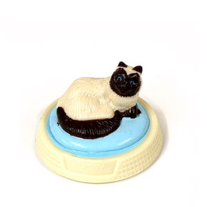 Op de afbeelding: Een beeldje van een Siamees met blauwe ogen, liggend op een lichtblauw kussen. Het kussen staat op een crèmekleurige basis met het woord "SASSY". De kat heeft een crèmekleurig lichaam en donkerbruine aftekeningen. De basis is ongeveer 5 cm in diameter.