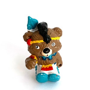 May include: A small, brown bear figurine dressed in a Native American style. The bear features a black mohawk, a blue feather, and a yellow headband. It wears a blue and white tunic and blue boots.