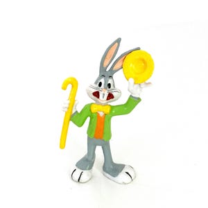 May include: A Bugs Bunny figurine, featuring the iconic cartoon character in a green jacket, orange vest, and yellow bow tie. He holds a yellow cane and a yellow disc. The figurine is gray, white, and red, with black accents.