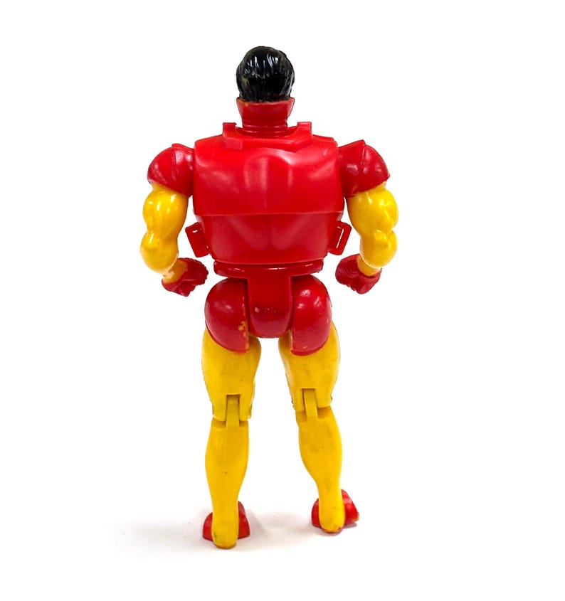 Puede incluir: Una figura de acci&oacute;n de pl&aacute;stico vista desde atr&aacute;s, con torso y brazos superiores rojos, antebrazos y piernas amarillas, y cabello negro. La figura tiene una complexi&oacute;n musculosa con articulaciones en hombros, caderas y rodillas.