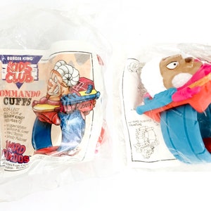 May include: Two Burger King Kids Club Commando Cuffs toys. One is blue, red, and orange with a character with a ram's head. The other is blue, red, and orange with a character with a brown head. The packaging says "Collect all four Burger King Kid's Club Commandos".