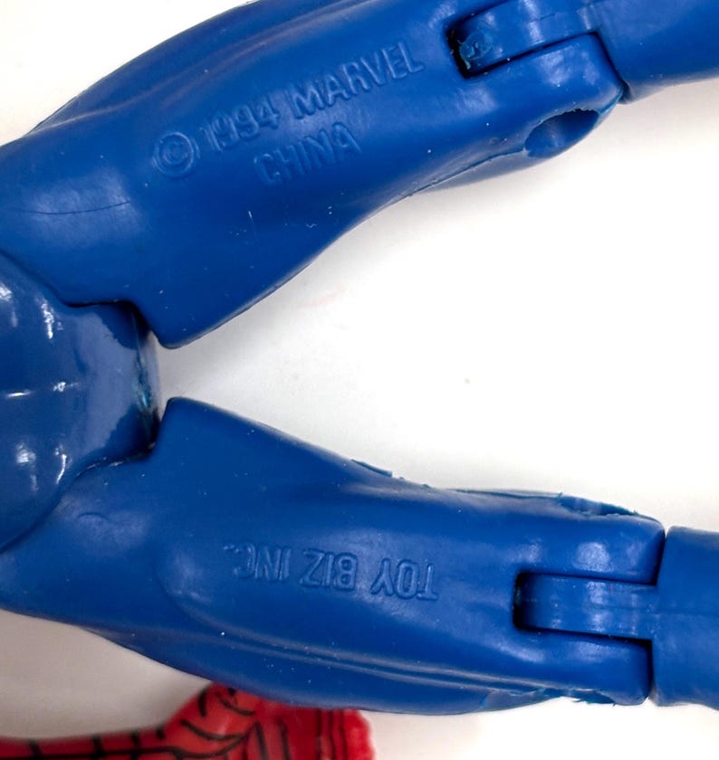 以下が含まれることがあります： 1994 MARVEL CHINAとTOY BIZ INC.という刻印がある青いプラスチック製のアクションフィギュアの脚部。関節部分があり、一部に赤と黒の模様が施された表面が見える。フィギュアのパーツは青いプラスチック素材で構成されている。