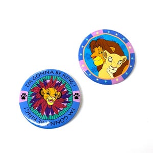Puede incluir: Dos botones redondos azules y rosas con imágenes de personajes de león de la película El Rey León. Un botón presenta a Simba con una melena roja y el texto "I'm gonna be king!" El otro botón presenta a Simba y Nala con el texto "I'm gonna be king!"