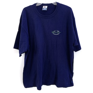 Puede incluir: Una camiseta azul marino con un logotipo blanco de Champion bordado en la parte delantera. El logotipo está rodeado por un círculo verde y negro.