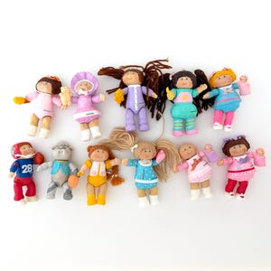 May include: A collection of ten small Cabbage Patch Kids plastic figurines featuring various outfits, hair colors, and accessories. The dolls are arranged in two rows, displaying diverse clothing styles including sports gear, pajamas, and dresses.