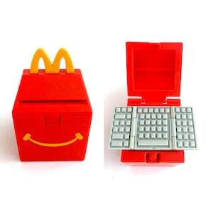 May include: A red plastic Happy Meal box with a yellow handle and a smiling face. Next to it is a red toy laptop with a gray keyboard, a fun collectible.
