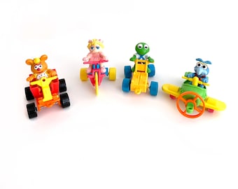 May include: Four Sesame Street character toys on wheels.  Miss Piggy is riding a pink tricycle, Kermit the Frog is driving a yellow ATV, Cookie Monster is driving a yellow airplane, and Big Bird is driving a red ATV.