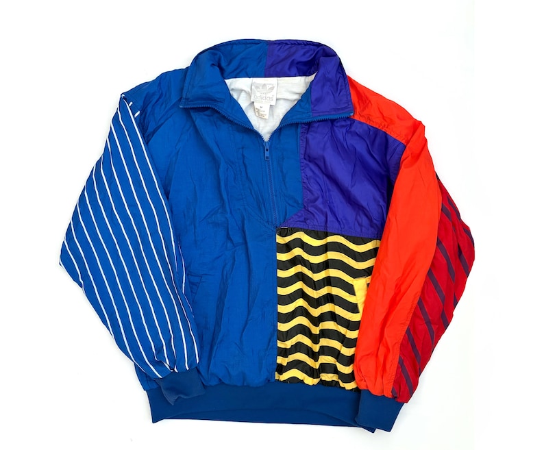 K&ouml;nnte beinhalten: Eine farblich abgesetzte Adidas-Pulloverjacke mit blauem Hauptteil, violettem Oberteil und rotem &Auml;rmel. Der linke &Auml;rmel hat wei&szlig;e vertikale Streifen, w&auml;hrend das untere rechte Feld ein gelb-schwarzes Wellenmuster aufweist. Sie hat einen hohen Kragen.