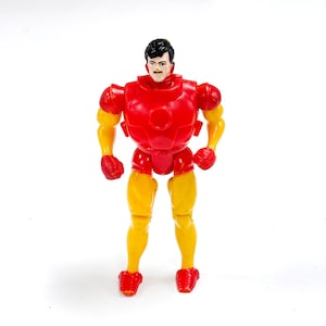 Puede incluir: Una figura de acci&oacute;n de pl&aacute;stico de un personaje con cabello negro y bigote, que viste un traje rojo y amarillo. La figura presenta una placa pectoral roja, hombreras rojas, guantes y botas rojas, con brazos y piernas amarillos. El personaje est&aacute; de pie en posici&oacute;n vertical con los brazos ligeramente doblados por los codos.