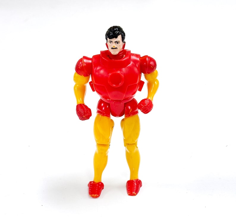 Puede incluir: Una figura de acci&oacute;n de pl&aacute;stico de un personaje con cabello negro y bigote, que viste un traje rojo y amarillo. La figura presenta una placa pectoral roja, hombreras rojas, guantes y botas rojas, con brazos y piernas amarillos. El personaje est&aacute; de pie en posici&oacute;n vertical con los brazos ligeramente doblados por los codos.