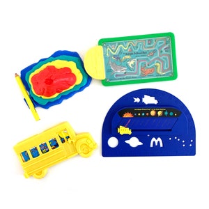 May include: A set of four plastic toys featuring a yellow school bus, a blue and yellow maze, a blue and yellow planet chart, and a red, yellow, blue, and green layered volcano. The toys are part of a McDonald's Happy Meal.