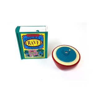 May include: A green and white "Pocket Travel Guide" book and a red and blue game piece. The book has the text "Carmen Sandiego" and "Pocket Travel Guide". The game piece has the text "Where in the world is Carmen Sandiego?"
