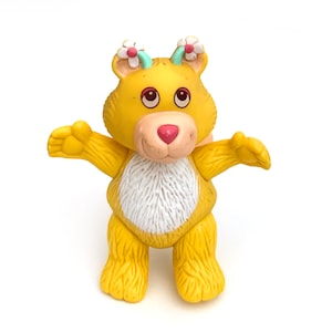 Op de afbeelding: Een gele berenfiguur met een witte borst, roze neus en bloemaccenten op het hoofd. De beer heeft zijn armen uitgestrekt en staat op een wit oppervlak. Het beeldje is een vintage speelgoed.