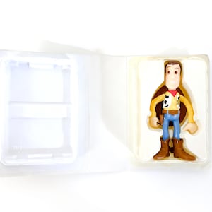 May include: A plastic toy figure of Woody from Toy Story. Woody is wearing his signature brown vest, yellow shirt, and blue jeans. He is holding his sheriff's badge and has a friendly expression on his face.
