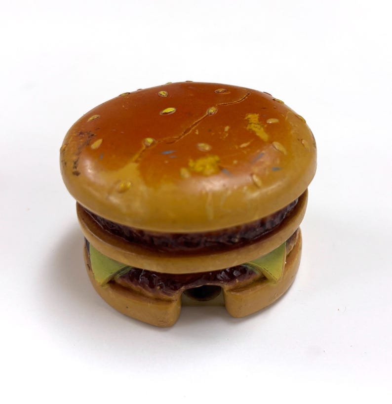 May include: A small, round, brown hamburger-shaped object with a glossy finish. The top bun features painted yellow sesame seeds. Visible layers include a dark brown patty, a light green slice of lettuce, and a tan bottom bun with a small cutout.