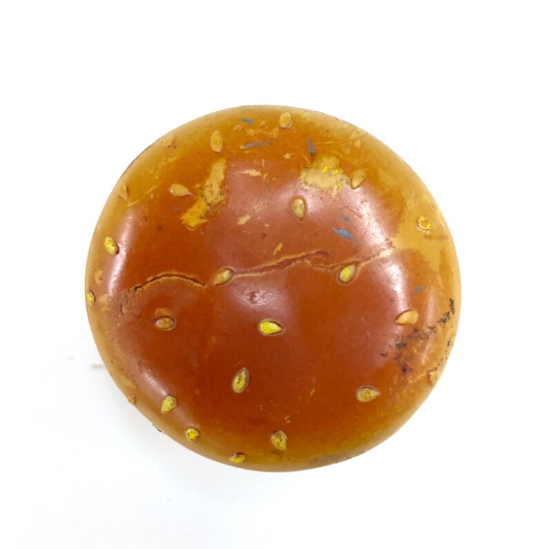 May include: A round, brown, glossy object resembling a hamburger bun, featuring a smooth, domed surface decorated with small, light yellow, seed-like shapes scattered across the top. The surface shows minor signs of wear and texture.