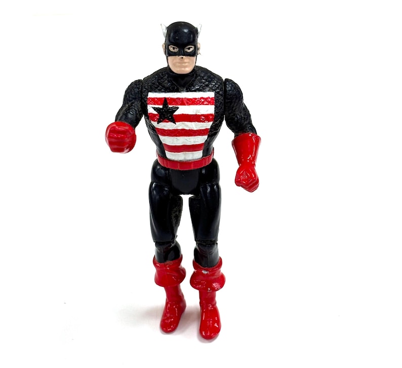Pu&ograve; includere: Un action figure in plastica di un personaggio che indossa una maschera e una tuta nera con un pannello pettorale a strisce rosse e bianche caratterizzato da una stella nera. Il personaggio ha guanti, cintura e stivali rossi, con arti articolati.
