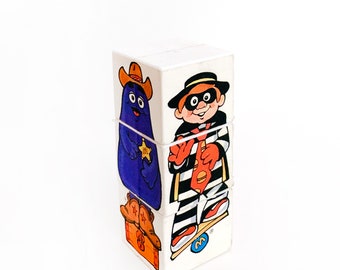 May include: A set of four white stacking blocks with colorful cartoon characters. The top block features a character wearing a black mask and striped shirt. The second block features a purple character wearing a cowboy hat. The third block features a character wearing a red and white striped shirt. The bottom block features a character wearing a cowboy hat and boots. The blocks are labeled with the letter "M".