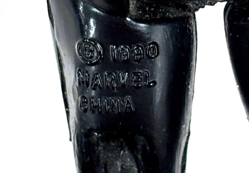 Pu&ograve; includere: Una gamba di action figure in plastica nera con testo stampato che recita &copy; 1990 MARVEL CHINA. La superficie &egrave; liscia e lucida, con le scritte in rilievo chiaramente visibili sul materiale scuro e solido del componente del giocattolo.