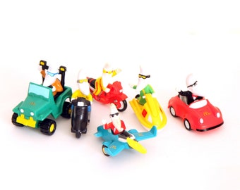 Pode incluir: Um conjunto de seis brinquedos de plástico com um personagem branco com cabeça de banana dirigindo diferentes veículos. Os veículos incluem um jipe verde, um scooter preto, uma motocicleta vermelha, um jet ski amarelo, um avião azul e um carro desportivo vermelho.
