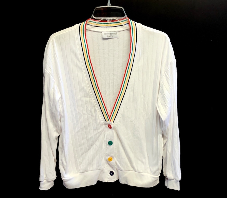 Pode incluir: Cardigan branco com gola listrada em vermelho, amarelo, verde e azul. O cardigan tem um decote em V e quatro bot&otilde;es coloridos.