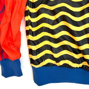 K&ouml;nnte beinhalten: Eine Jacke mit einem horizontalen, wellenf&ouml;rmigen Streifenmuster in Schwarz und Gelb auf dem Hauptteil, kombiniert mit einfarbig roten &Auml;rmeln sowie einem blauen Rippstrickb&uuml;ndchen am Saum und an den &Auml;rmeln. Das Material ist glatt und leicht.