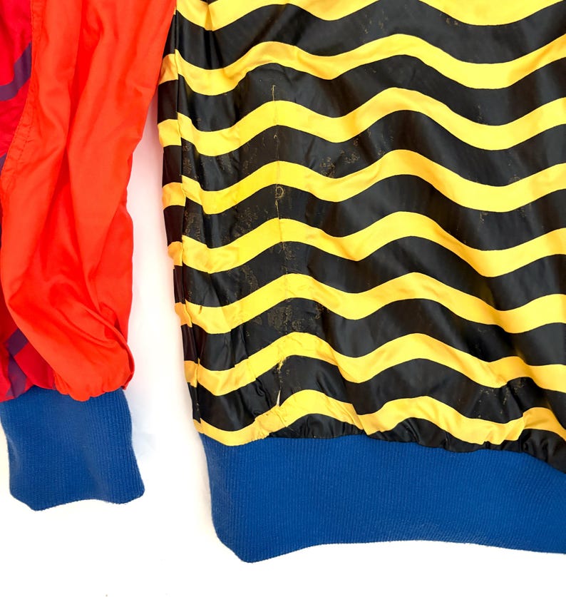 K&ouml;nnte beinhalten: Eine Jacke mit einem horizontalen, wellenf&ouml;rmigen Streifenmuster in Schwarz und Gelb auf dem Hauptteil, kombiniert mit einfarbig roten &Auml;rmeln sowie einem blauen Rippstrickb&uuml;ndchen am Saum und an den &Auml;rmeln. Das Material ist glatt und leicht.