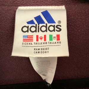 K&ouml;nnte beinhalten: Ein wei&szlig;es Etikett mit dem Adidas-Logo und dem Text "adidas" in Schwarz. Das Etikett enth&auml;lt auch den Text "SIZEXL TAILLE XG TALLAXG", "RN# 88387" und "CA# 00411". Das Etikett hat eine amerikanische Flagge in Rot, Wei&szlig; und Blau, eine kanadische Flagge und eine mexikanische Flagge.
